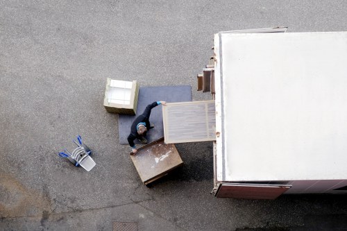 An aerial view of a person in casual clothing working outdoors on a residential driveway, surrounded by various household items and moving equipment. The individual is handling a light-colored wooden side table with a flat top and vertical slats on the sides, positioning it either onto or off a large, white moving van with its rear doors open. Nearby, there are multiple cardboard boxes, one of which is brown and slightly worn, and another gray fabric-covered cushion or small piece of furniture. To the left, a blue and white tool or packing strap is laid on the ground, along with a white plastic container. The scene suggests a house removal or moving process taking place in Bermondsey, with the background showing part of the residential street, indicating a typical house move environment. The lighting appears natural, possibly during daylight hours, contributing to a clear view of the items and activity involved.